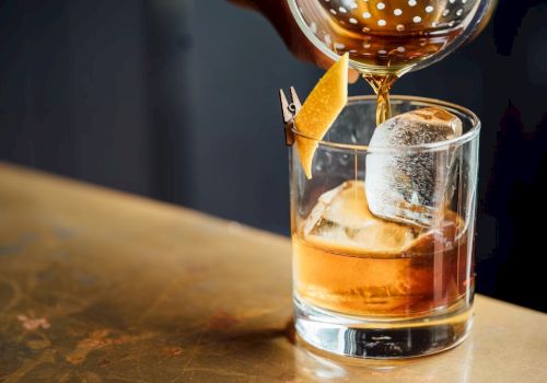 A glass with whiskey and ice on a bar, garnished with an orange peel, a large ice cube, and a pour of another drink in a copper-topped shaker.