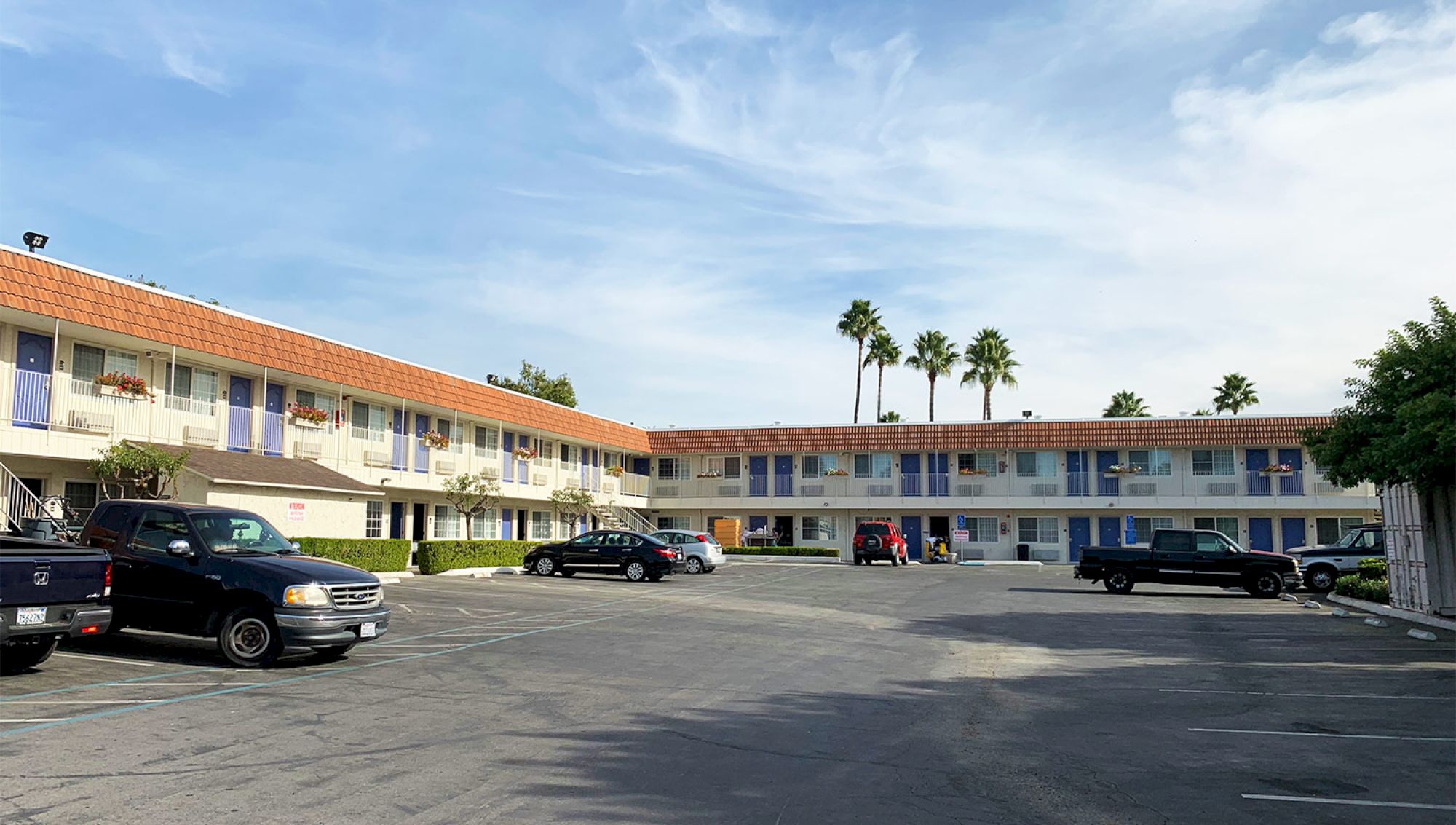 A two-story motel-style building with orange-tiled roof, blue doors/windows, a central parking lot, several parked cars, and palm trees in the background.