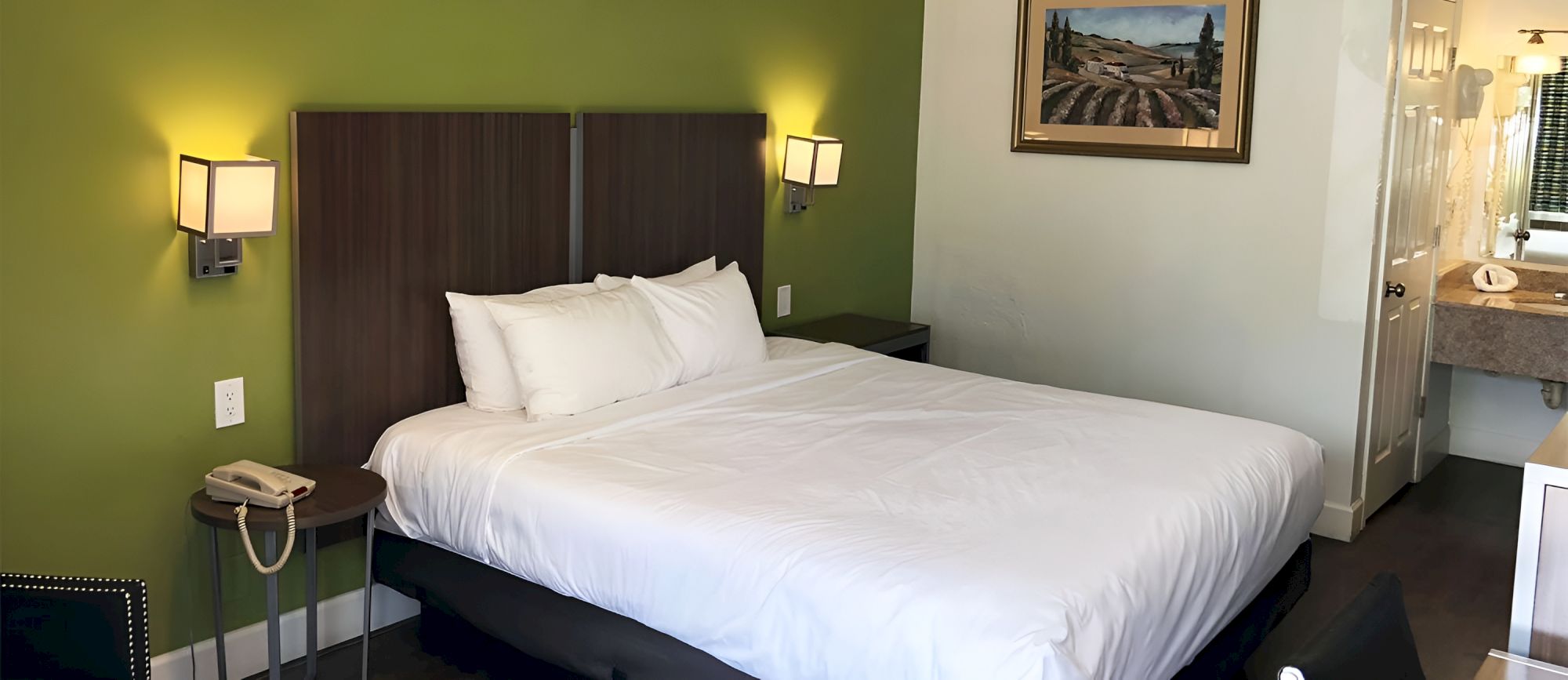 A tidy hotel room with a large white bed against a lime-green wall, two wall lamps, a small side table with a phone, and a view into a bathroom.
