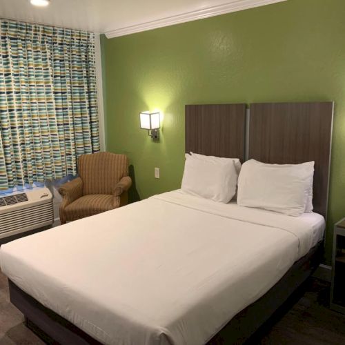 A hotel room with a green wall, queen bed, two nightstands, wall lamps, a patterned curtain, a chair, and a window AC unit.