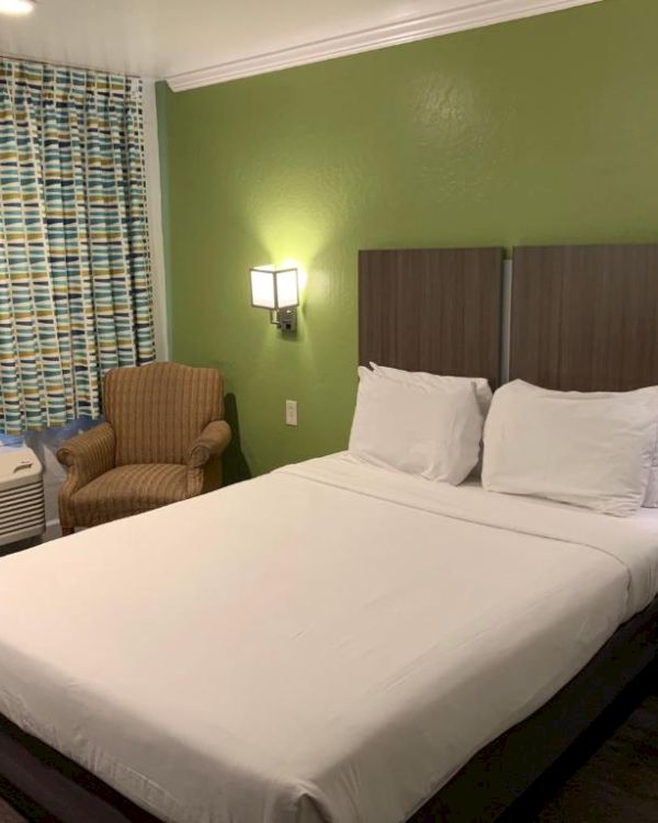 A hotel room with a green wall, queen bed, two nightstands, wall lamps, a patterned curtain, a chair, and a window AC unit.