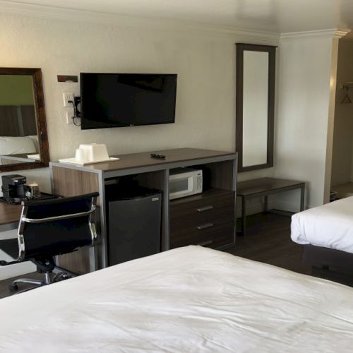 A hotel room with two beds, a desk and chair, wall-mounted TV, mirror, mini-fridge, microwave, and a small kitchenette area near an open doorway.