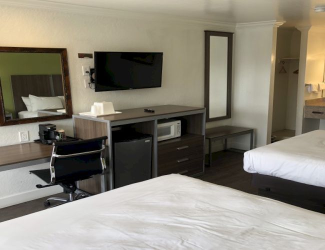 A hotel room with two beds, a desk and chair, wall-mounted TV, mirror, mini-fridge, microwave, and a small kitchenette area near an open doorway.