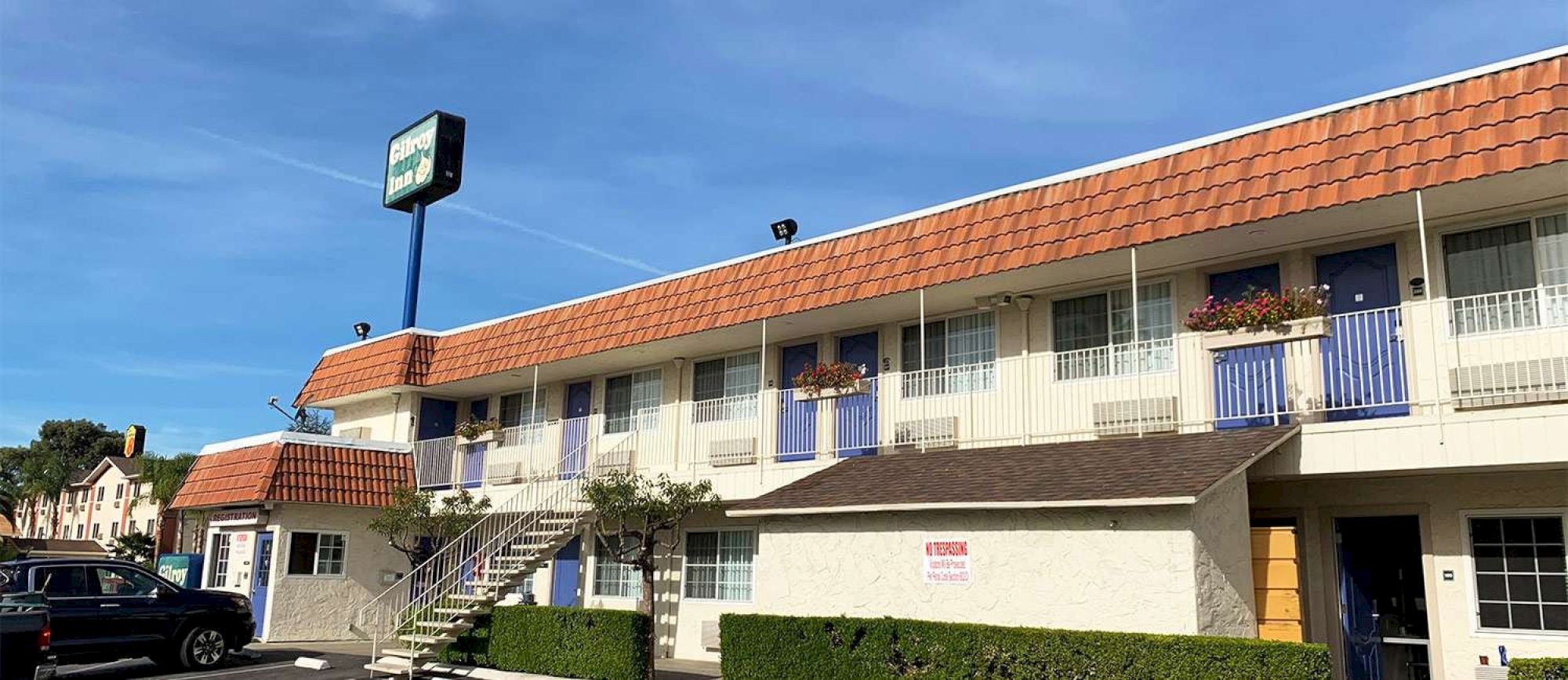A two-story motel with blue doors, orange-tiled roof, small balconies, and a sign on a blue pole; cars parked outside under a sunny sky.
