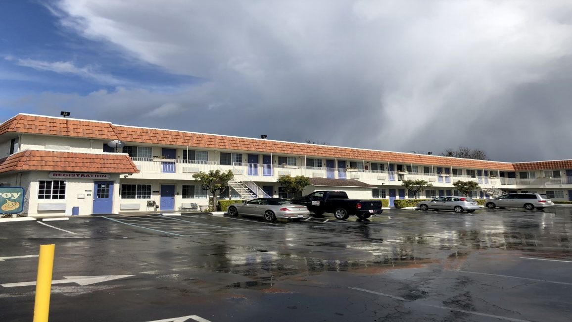 A single-story motel with a long row of blue doors, a red-tiled roof, a wet parking lot, and a cloudy sky overhead, with few cars parked.