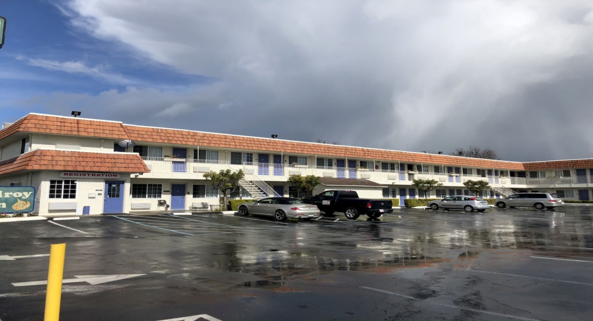 A single-story motel with a long row of blue doors, a red-tiled roof, a wet parking lot, and a cloudy sky overhead, with few cars parked.