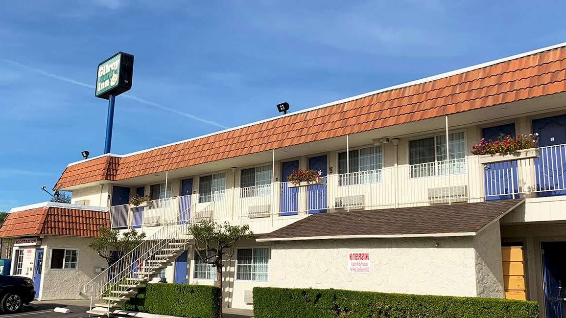 A two-story motel with a tan, orange-tiled roof, blue doors, and staircases outside each unit; sign on a blue pole against a sunny sky.