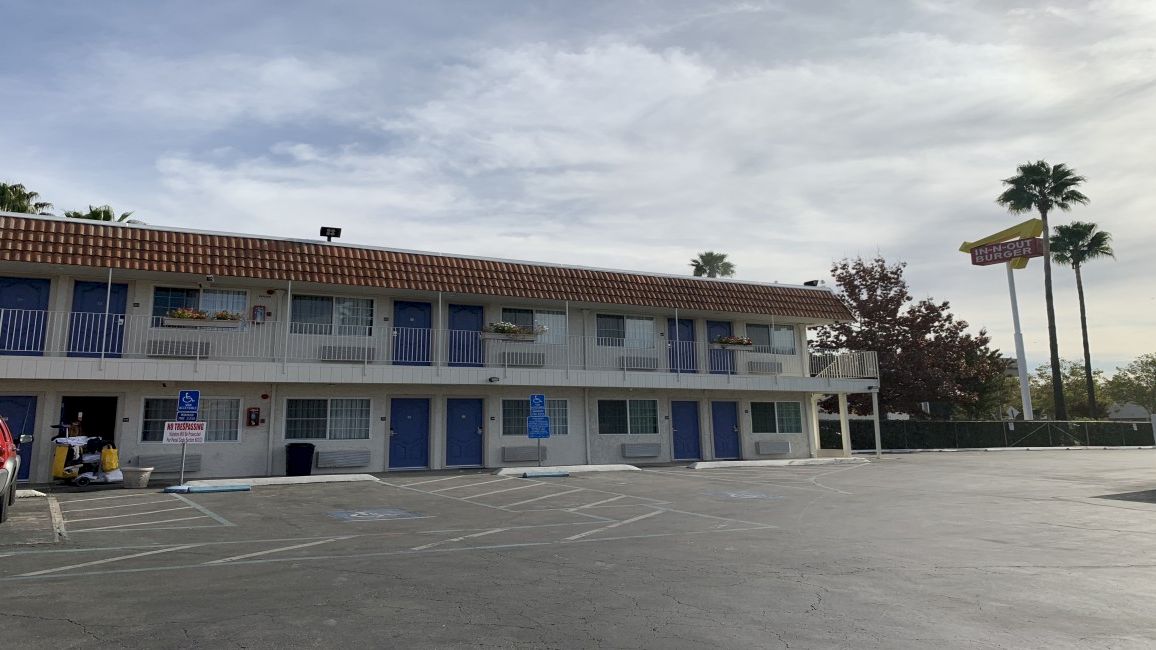 A two-story motel with blue doors and white trim, a large empty parking lot, and a few palm trees against a partly cloudy sky.