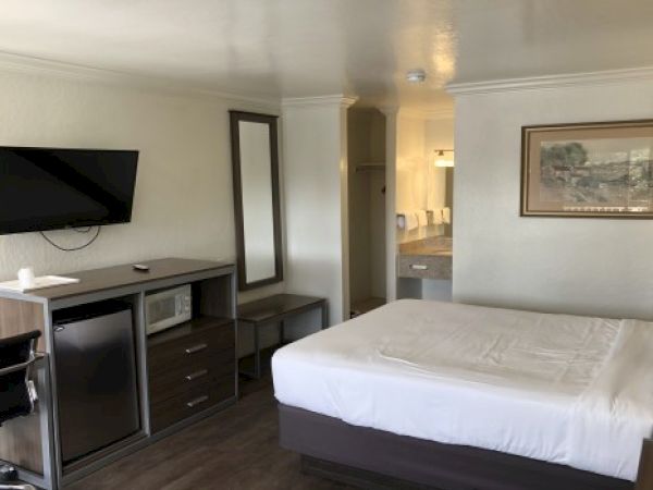 A hotel room with a queen bed, a wall-mounted TV, a desk with a chair, a dresser with a mirror, and a framed picture on the wall, plus a view into the bathroom.