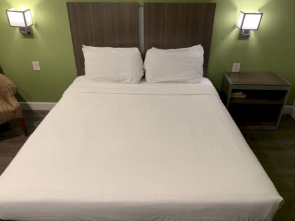 A neatly made bed with two pillows on a green-walled hotel room, bedside tables, and wall lamps. Top it at 140 characters, always ending the sentence.