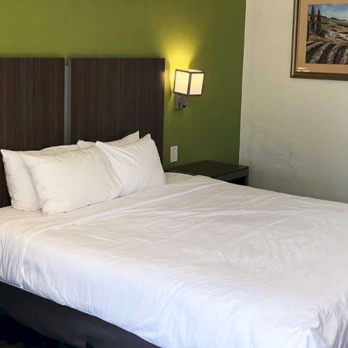 A hotel room with a large white bed against a green accent wall, two wall lamps, a bedside table with a phone, and a small seating area, plus a doorway to a bathroom.