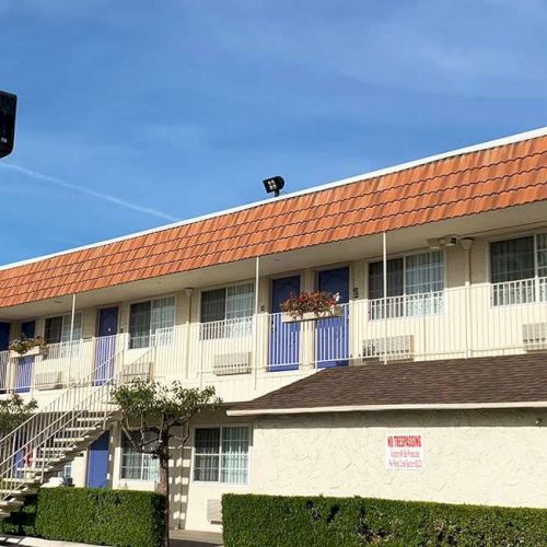 A two-story motel with a red tiled roof, blue doors, white railings, and a sign on a tall pole under a blue sky, with parked cars nearby.