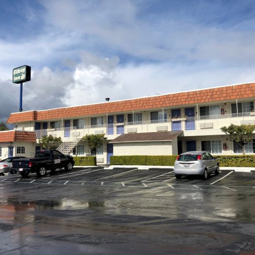 A two-story motel with a red-tile roof, a tall sign, palm trees, and a wet parking lot with a few cars.