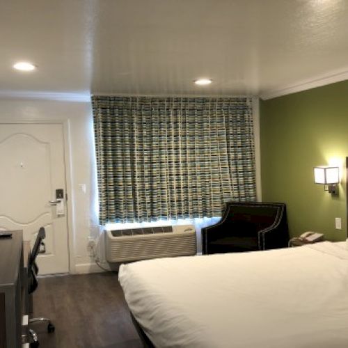 A hotel room with a large bed, green accent wall, desk and chair, flat-screen TV, window with patterned curtains, and a small seating area by the window, all lit softly.