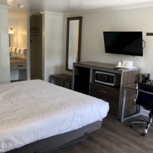 A modern hotel room with a large bed, desk, mounted TV, a desk chair, a microwave cabinet, a mirror, and a doorway to a small bathroom/entrance.