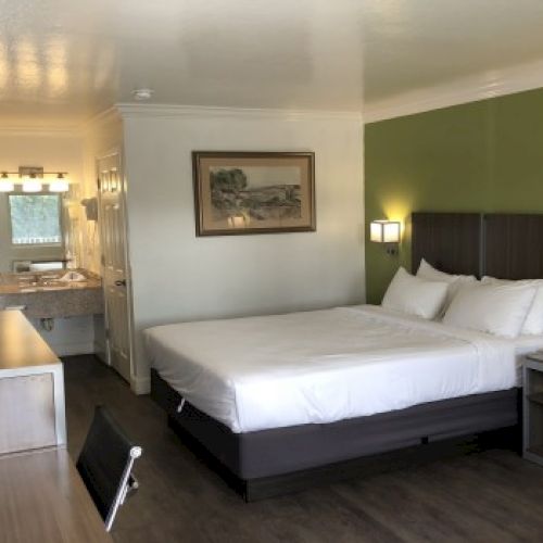 A modern hotel room with a large bed against a green accent wall, a desk, flat-screen TV, and an open bathroom visible in the background.