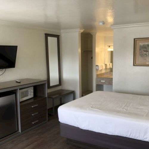 A hotel room with a bed, desk, mounted TV, mini-fridge, microwave, mirror, framed artwork, and a view toward an open kitchenette and hallway.