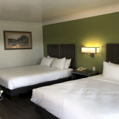 A hotel room with two double beds, white linens, a green accent wall, wall lamps, a framed painting, and a small desk area by the door.