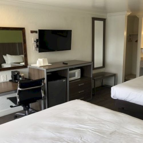 A hotel room with two beds, a desk and chair, mounted TV, mirror, mini-fridge, coffee maker, and a doorway to a small bathroom area.