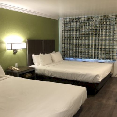 A hotel room with two double beds, a nightstand, wall lamp, green accent wall, patterned curtains, desk and chair near the door.