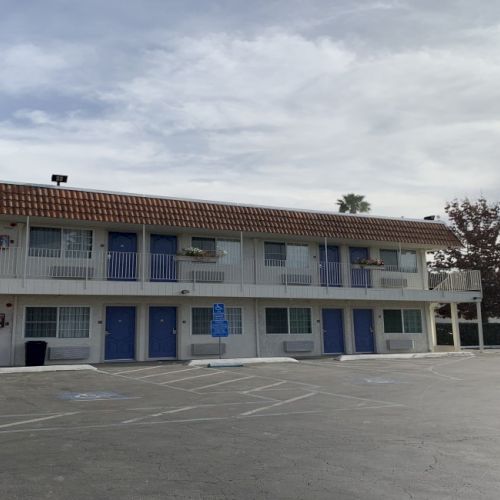A two-story motel with blue doors and white trim, a mostly empty lot, palm trees, and a sign on the right under a cloudy sky.