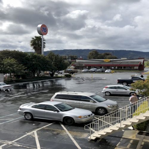 A rainy parking lot outside a motel or roadside stop, several parked cars, damp pavement, a stairway on the right, and rolling hills in the distance.