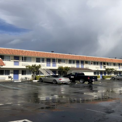 A row of two-story motel rooms with blue doors, a wet parking lot, several cars, and a cloudy sky overhead, in a seaside motel setting.