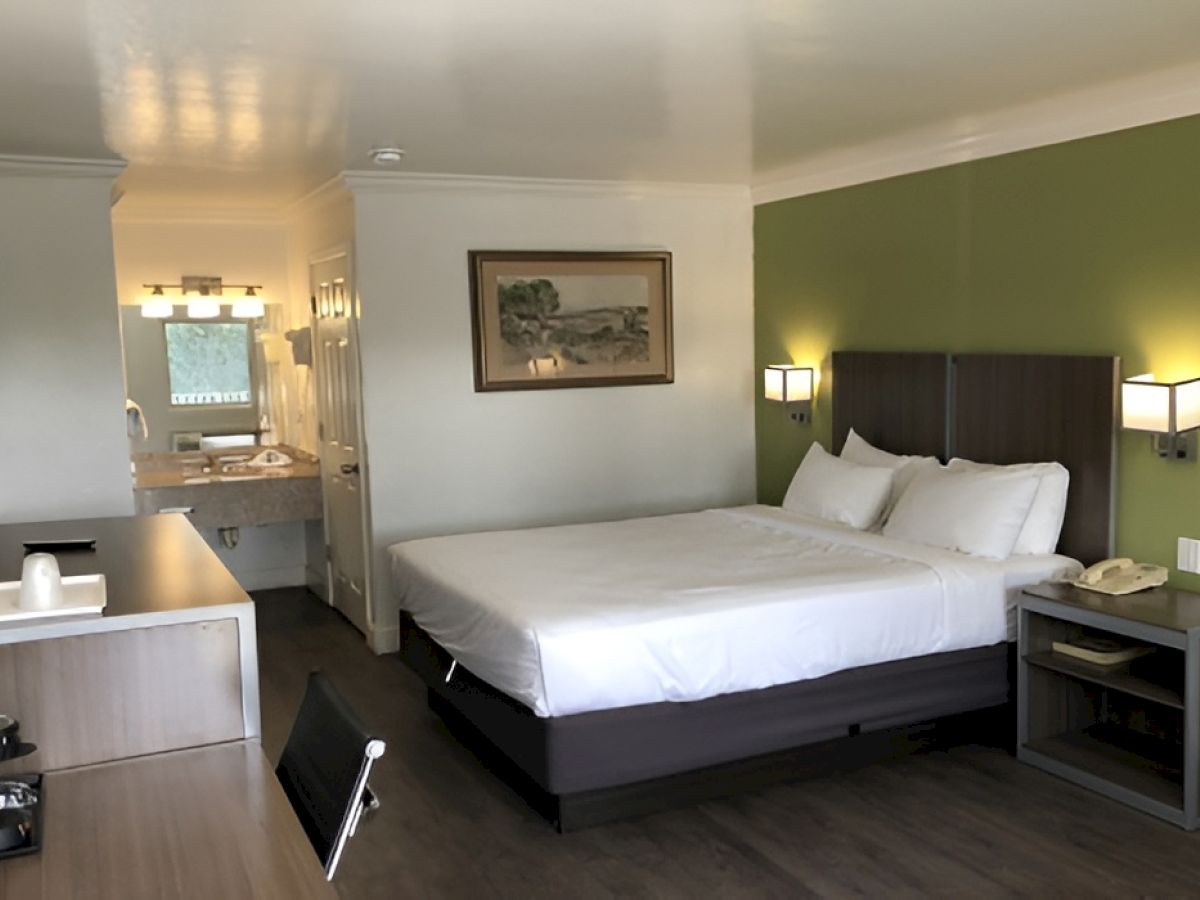 A modern hotel room with a king bed, green accent wall, two nightstands, a desk, and a small bathroom area visible in the back, stylish and tidy.