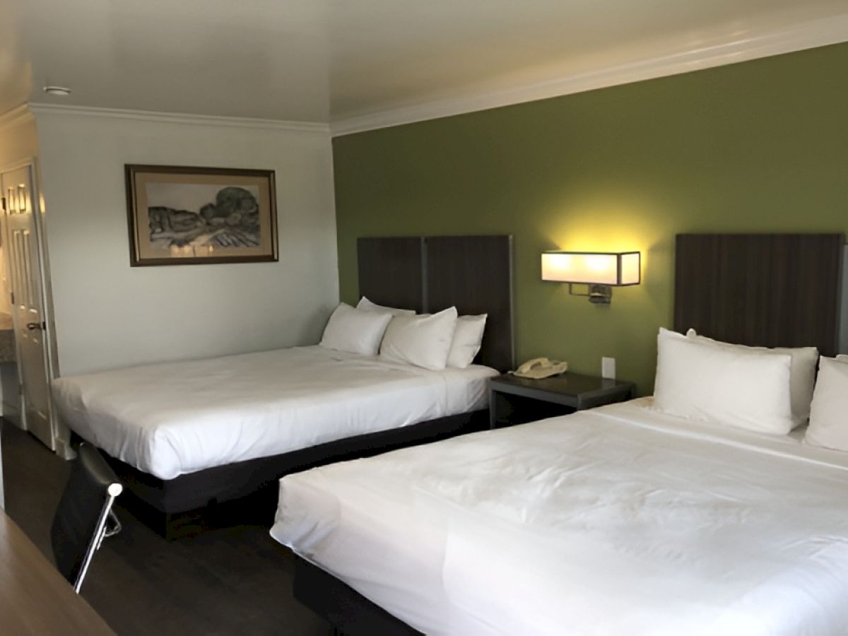 A hotel room with two large beds, green accent wall, nightstands, wall lamp, and a framed picture on the wall, plus a bathroom area in the background.