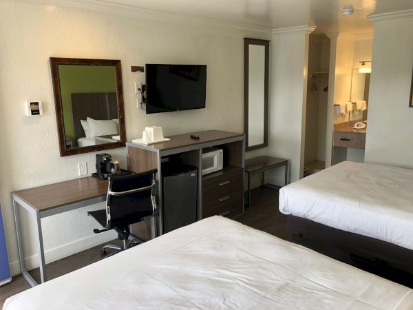 A tidy hotel room with two beds, a desk, chair, wall-mounted TV, mirror, mini-fridge, and a small seating area near a doorway.