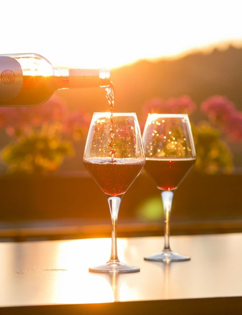 Two wine glasses on a table, sunset glow, bottle pouring red wine into one glass, outdoor vineyard backdrop, warm and romantic.