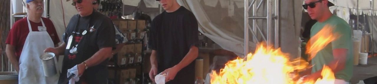 Chefs in hats and aprons cook over blazing open flames at an outdoor event, with several people working near the fire.